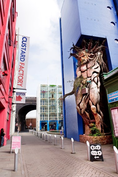 View along pedestrianised street with Green Man sculpture on the right, factory buildings either side and railway arch in the distance.