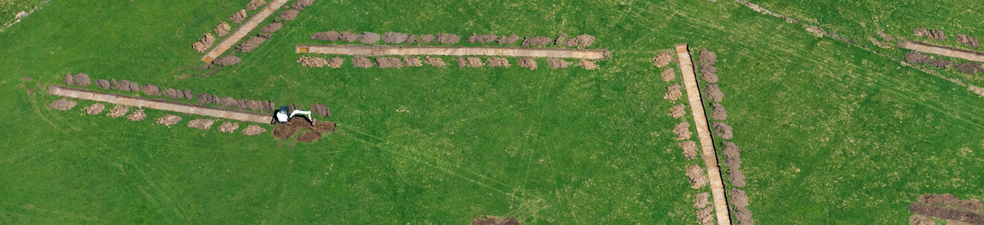 An aerial photograph of eight trial archaeological trenches in a green field.