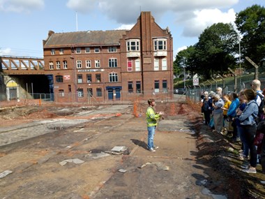 A photograph of people listening to a talk at an archaeological excavation site at Angel Meadow.