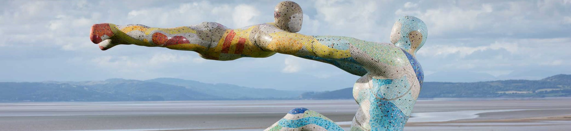 A sculptural installation in a coastal bay setting, depicting a seated female figure swinging a child in mid-air.