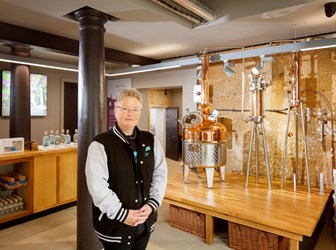 A short-haired lady standing with her hands clasped in front of a gin distillery. 