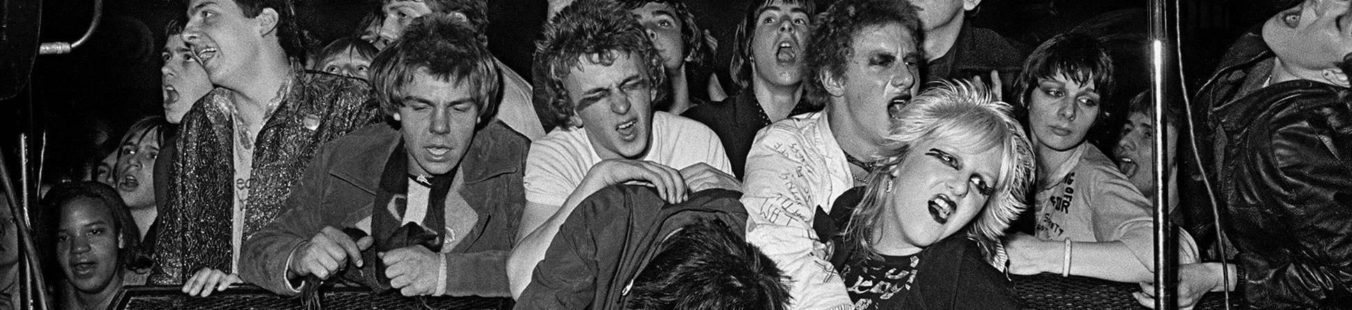 Punk girl in fishnets and suspenders reclining between two monitors up on stage with other members of the gig audience behind and around her. Photo taken 1976.