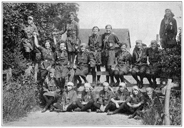 A group of girls in girl guide uniforms sitting on a wooden fence and on the ground.