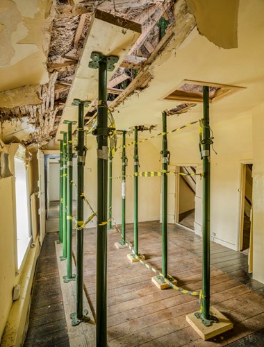 A series of metal poles standing on timber supports braced between the floor and a damaged ceiling.